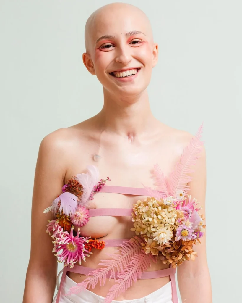 Donna sorridente con testa rasata e composizione floreale rosa sul busto durante uno shooting del progetto Feeling Nova, dedicato alla rinascita e alla riconnessione delle donne con il proprio corpo.