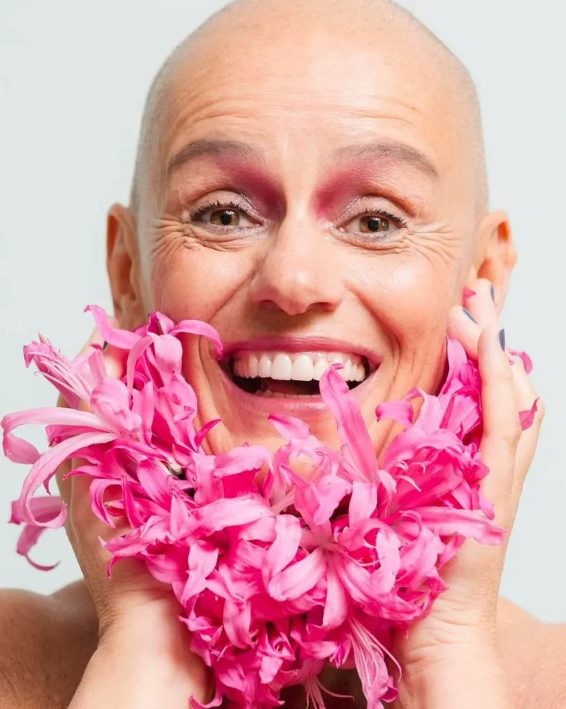 Ritratto di donna con testa rasata e fiori rosa durante uno shooting di Feeling Nova, progetto che promuove consapevolezza, rinascita e relazione con il proprio corpo.